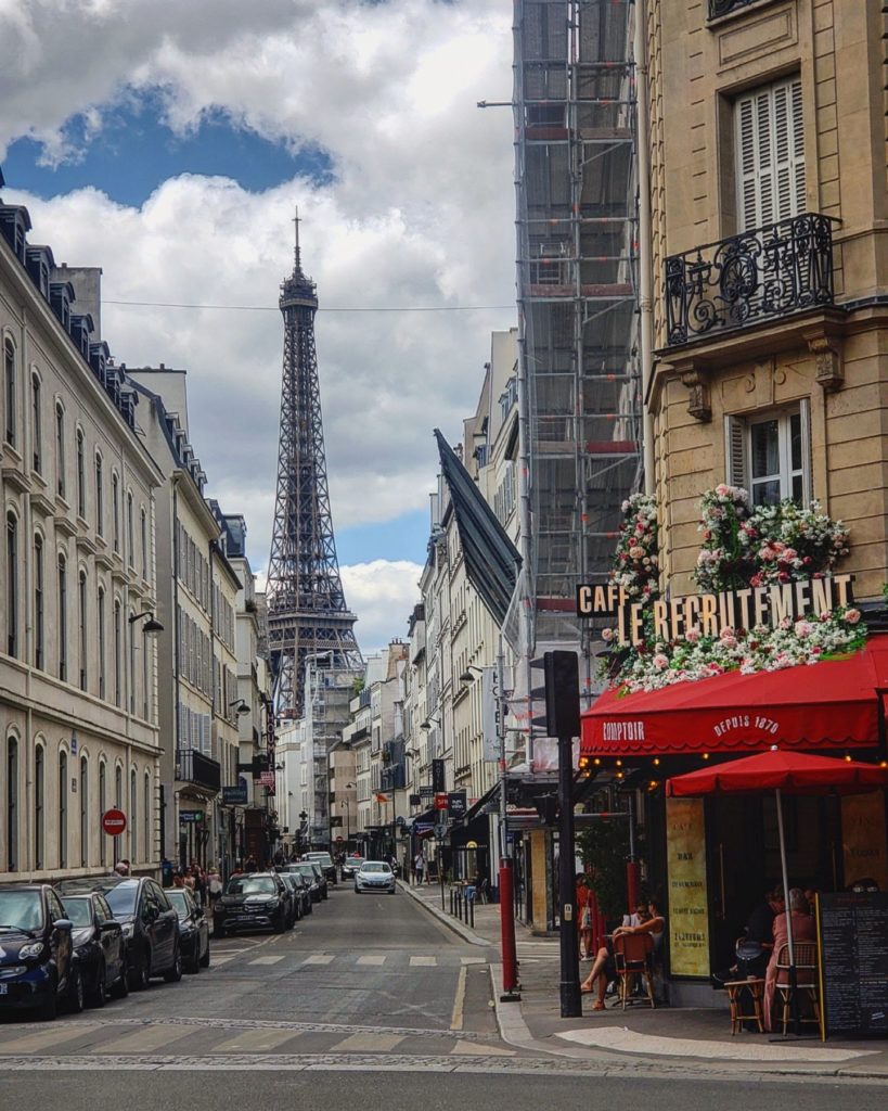 View of the Eiffel Tower hiding between two typical Parisian buildings, both sides of the street are packed with parked cars, with a view of a famous cafe, le Recruitment, on a street corner. There is a red awning that covers a patio and there are fake flowers that are poking out from the cafe's sign.